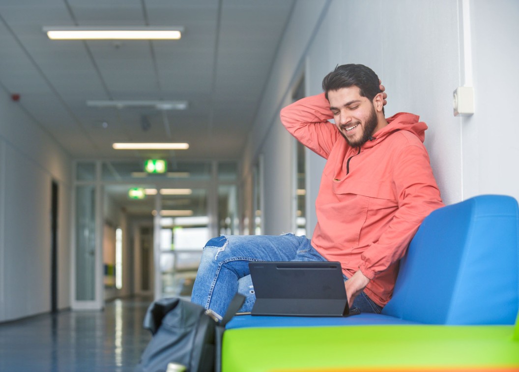 Studierender sitzt auf dem Flur der Hochschule und schaut auf ein Tablet.