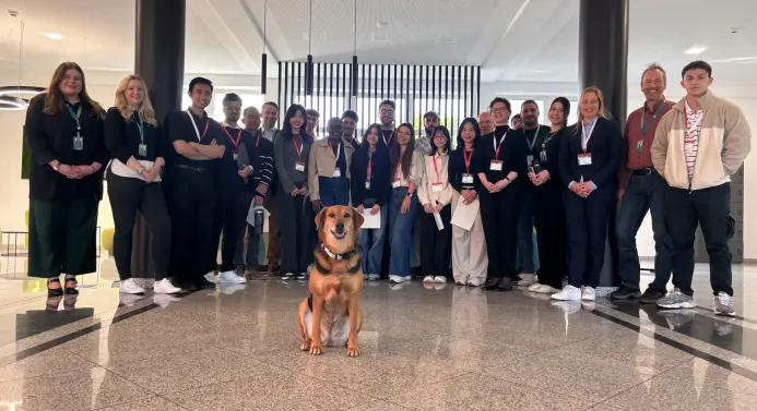 Eine Gruppe von Menschen steht in einem Halbkreis, davor sitzt ein Hund.