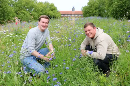 Zwei junge Studenten hocken auf einer Blühwiese und lächeln in die Kamera
